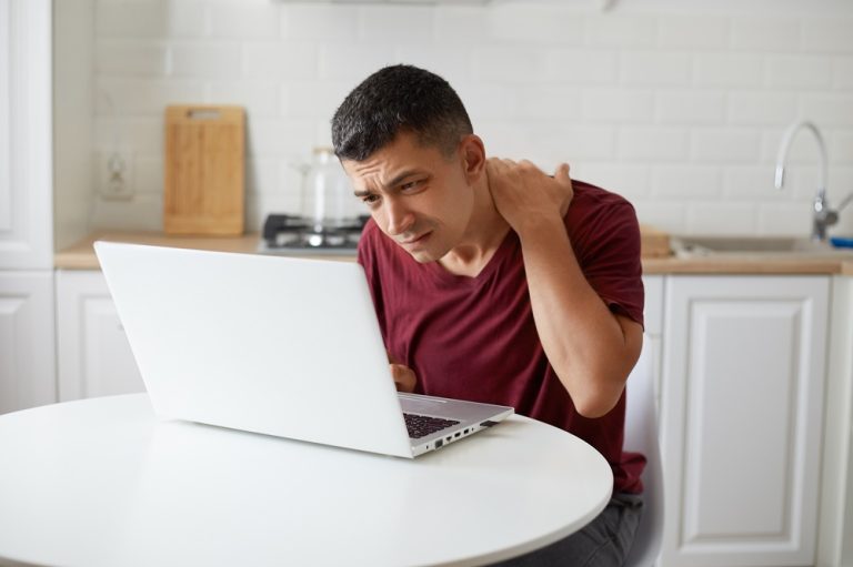 Indoor shot of freelancer male worker sitting in kithen at table in front of white notebook, looking intently at laptop display trying to notice importaint thing, keeps hand on neck